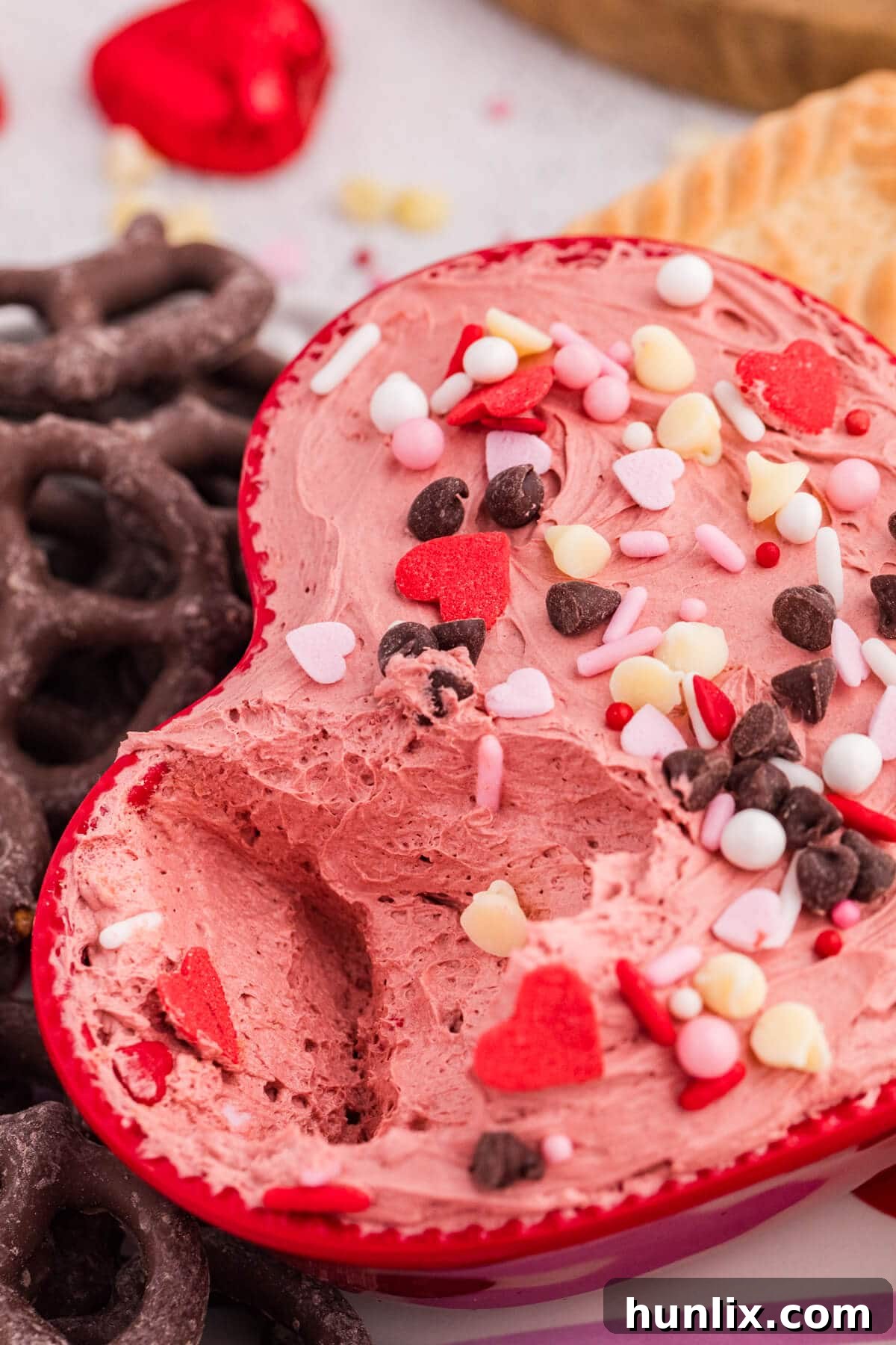 Scooped pink cheesecake dip in a red heart-shaped bowl topped with Valentine’s sprinkles and chocolate chips.