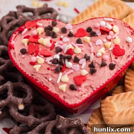 Heart-shaped bowl of Valentine’s cheesecake dip with sprinkles, paired with pretzels and rectangular cookies on a dessert platter.