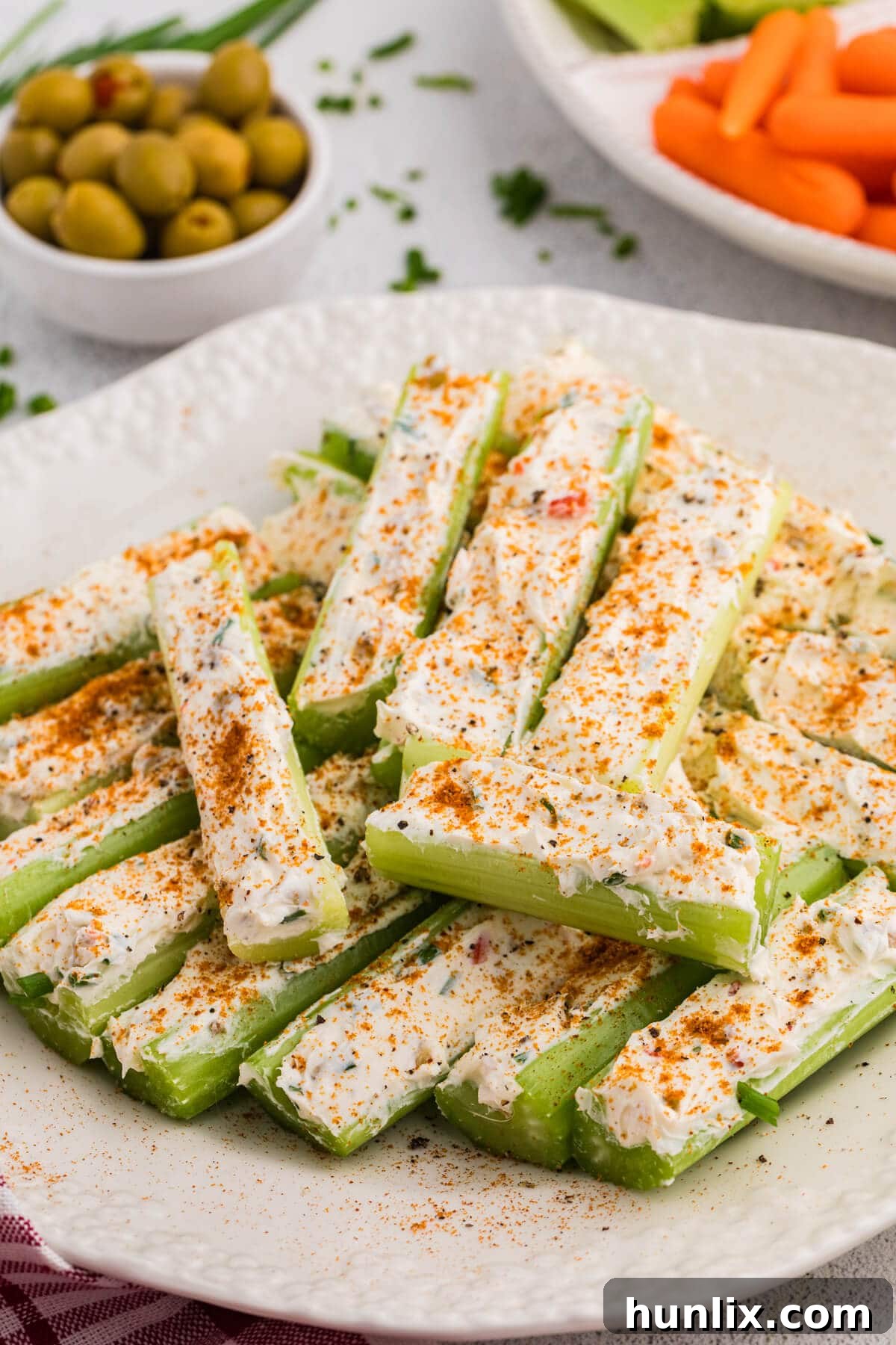 Fresh celery sticks filled with creamy herb cheese spread and sprinkled with paprika on a white plate, served with olives, carrot sticks, and celery pieces on a light tabletop.