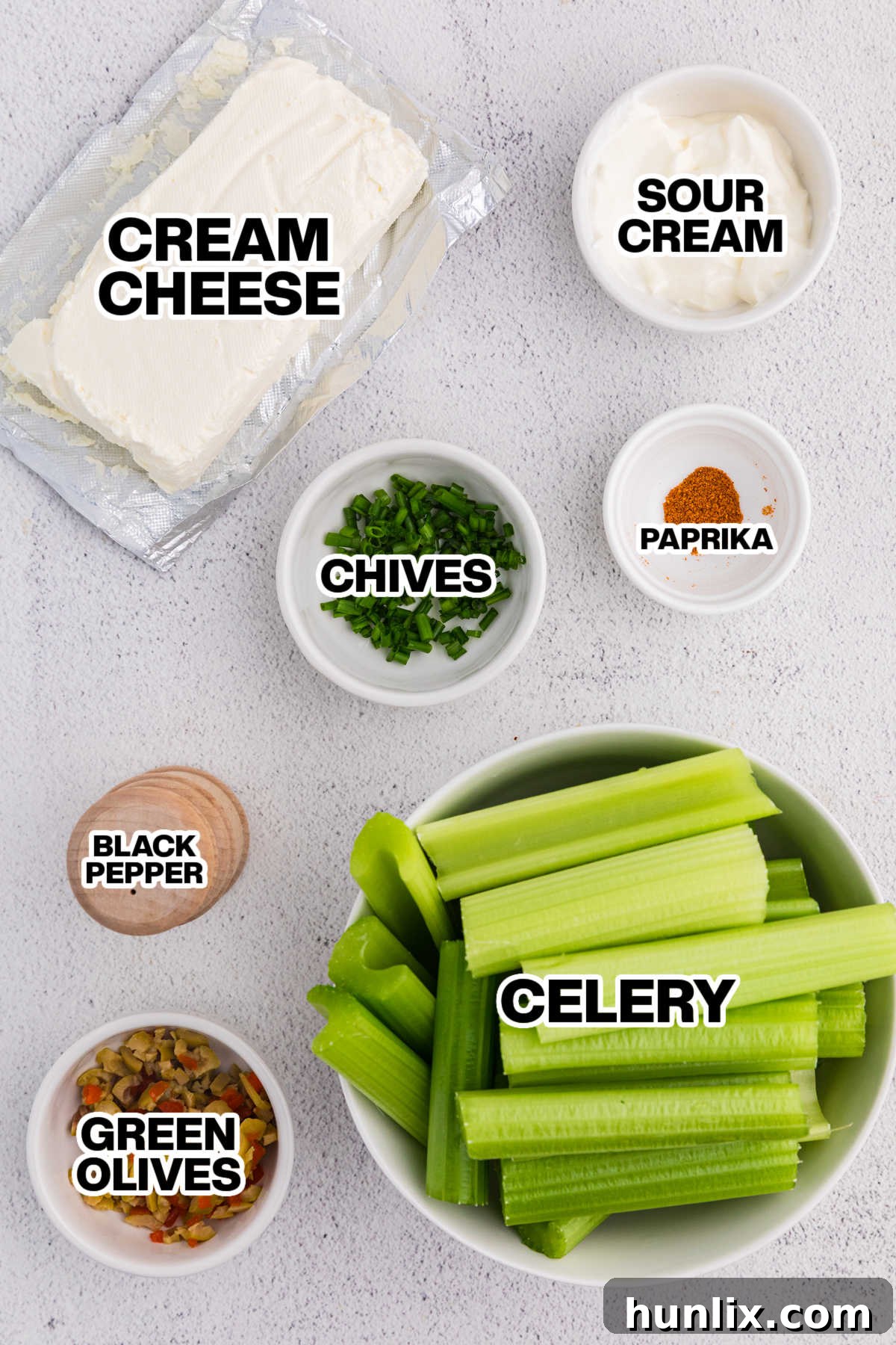 Ingredients for stuffed celery laid out on a white background, including celery stalks, cream cheese, sour cream, green olives, chives, and paprika.