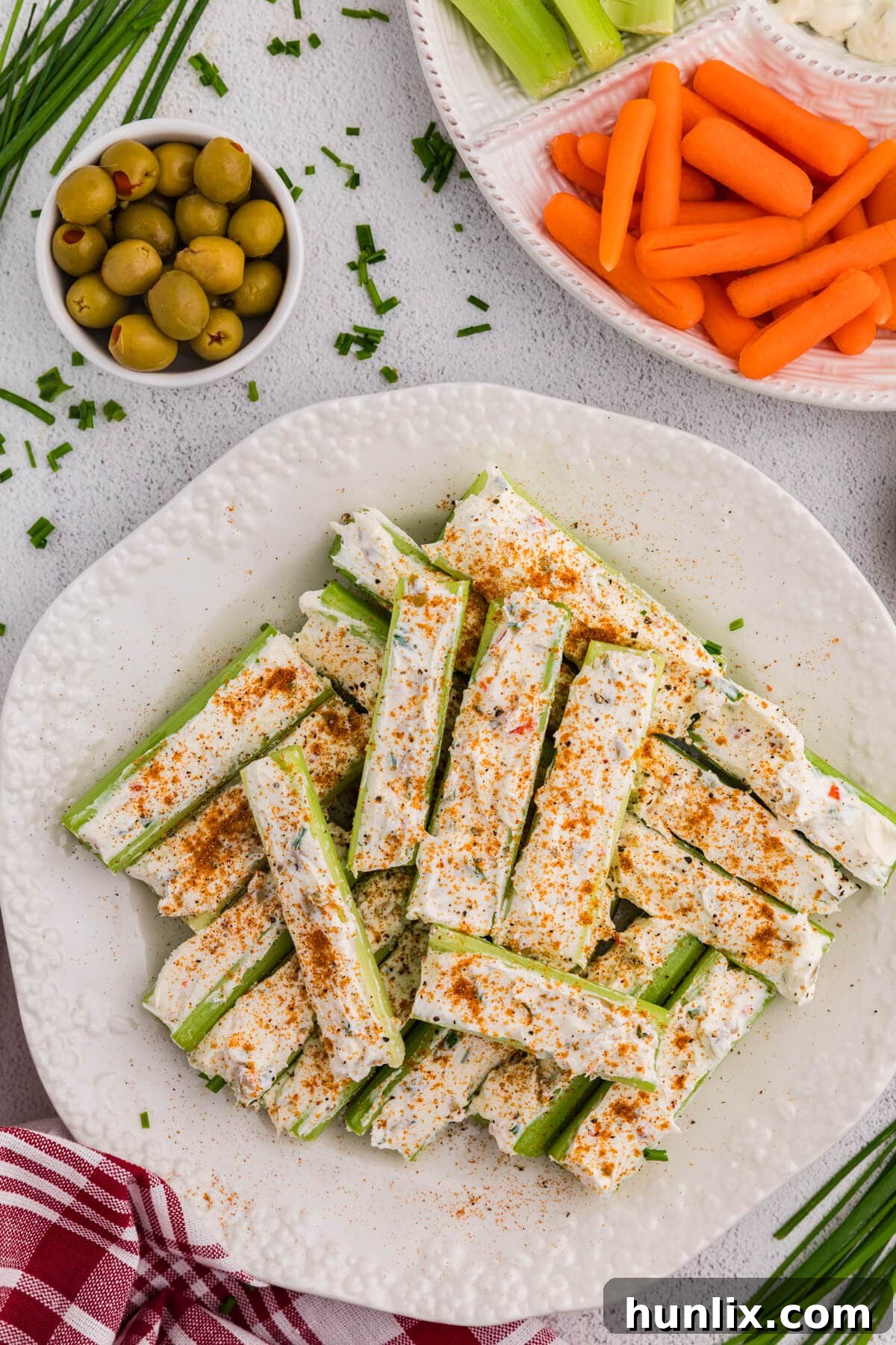 Celery sticks stuffed with a creamy herb cheese mixture and topped with paprika and black pepper, plated on a white dish with olives, carrots, and chopped chives in the background.