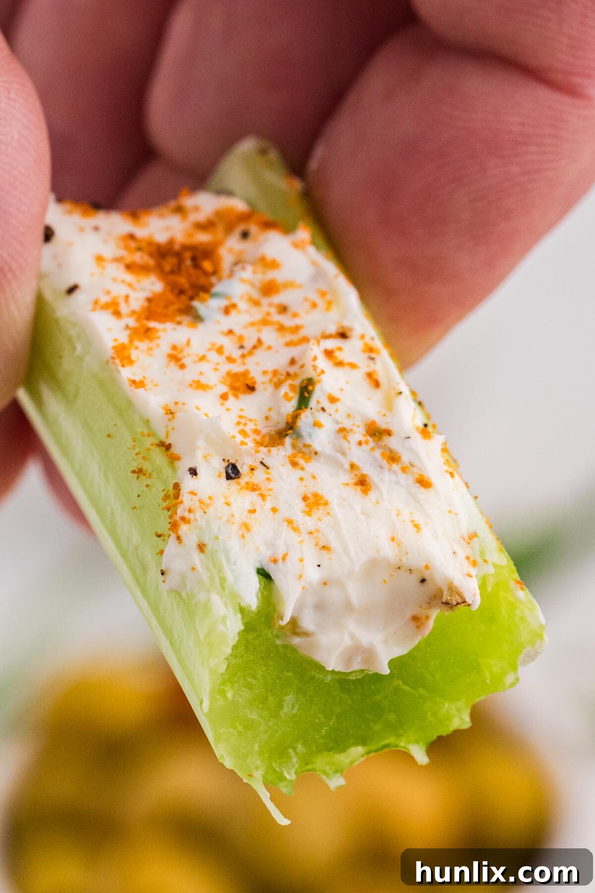 Macro view of a hand holding a celery stick stuffed with creamy herb cheese and sprinkled with paprika, showing the crunchy celery texture and smooth filling.