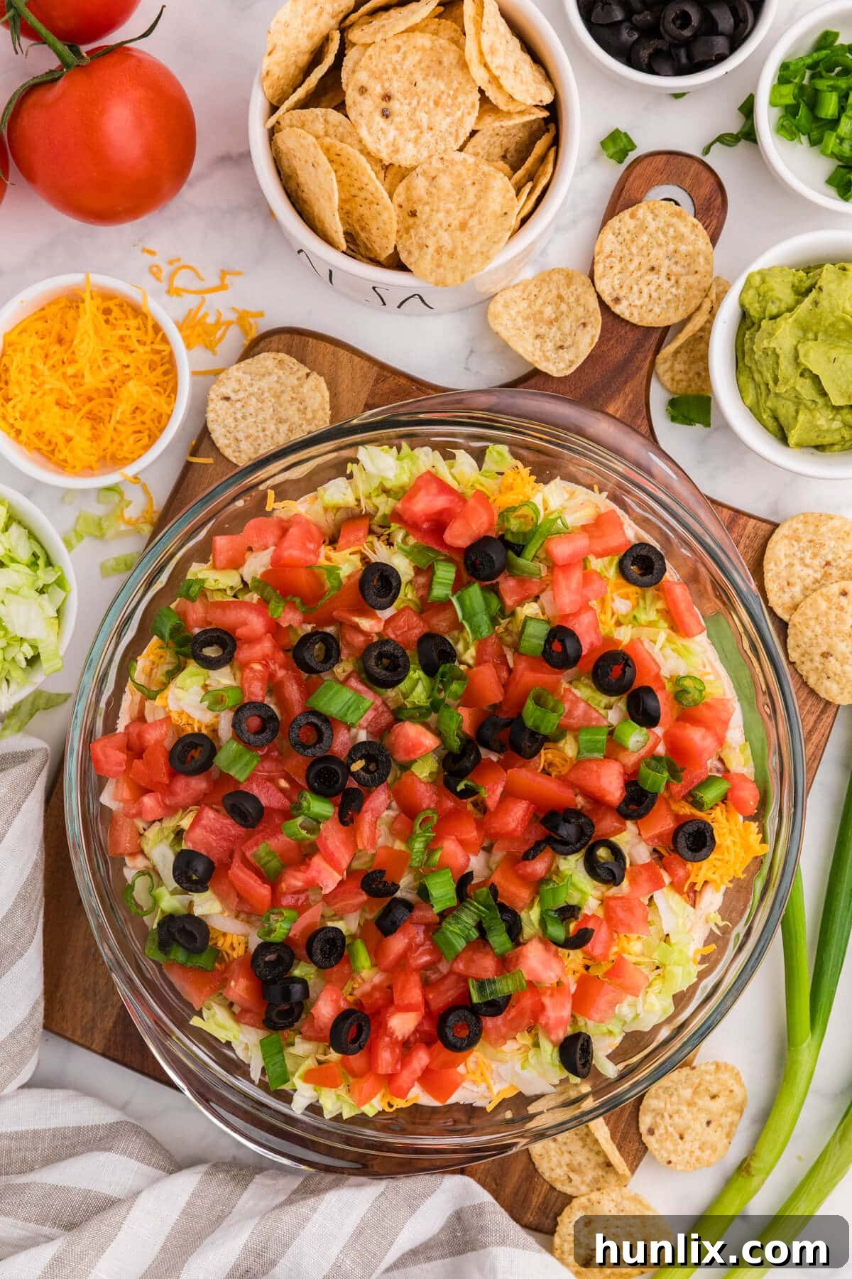 Taco Dip layered appetizer in glass dish topped with diced tomatoes, black olives, green onions, shredded lettuce and cheddar cheese, surrounded by tortilla chips, guacamole, salsa, and fresh ingredients on marble countertop.