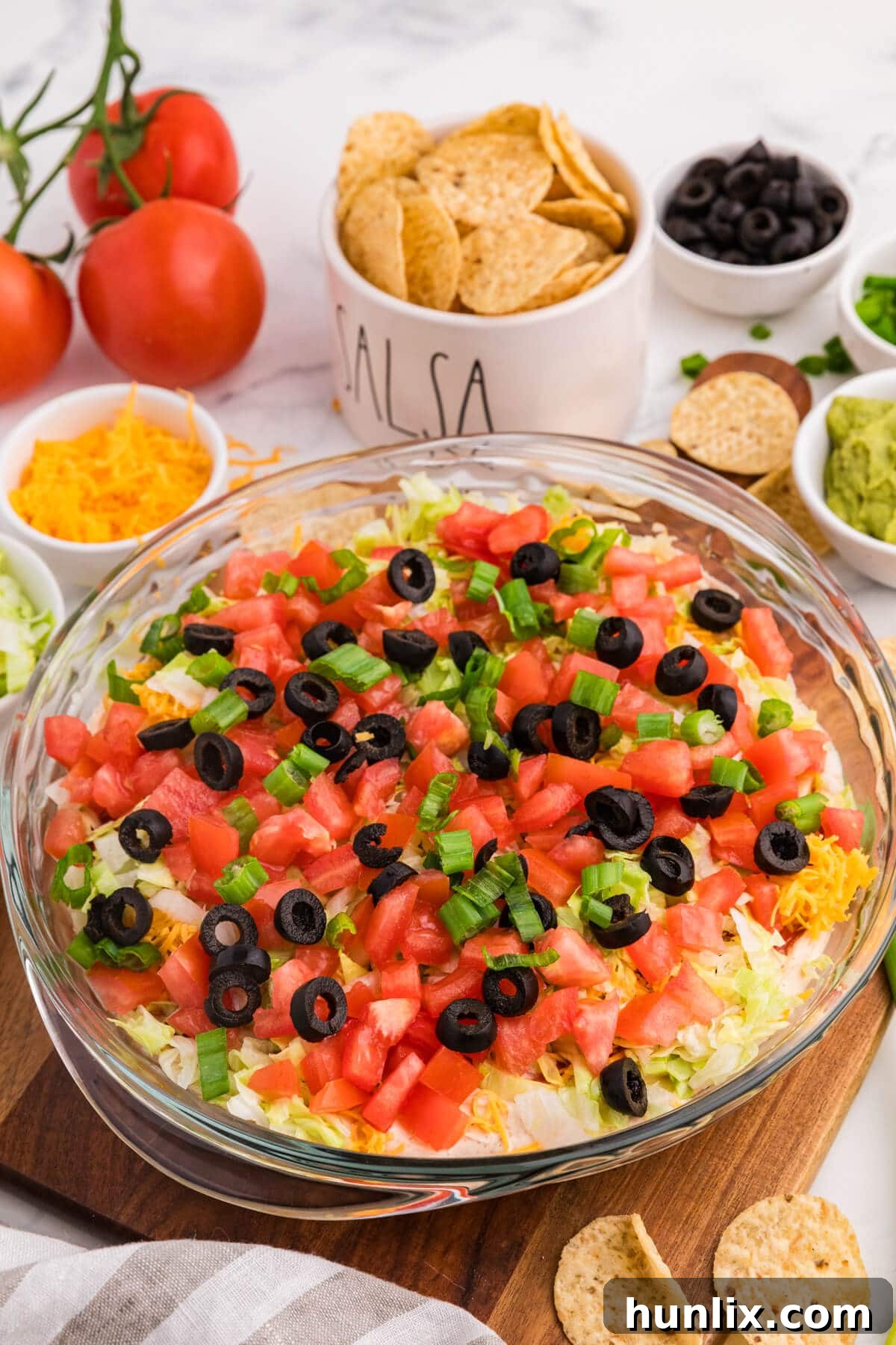Side view of creamy taco dip layered with beans, sour cream, cheese, lettuce, tomatoes, olives, and green onions in glass dish on wooden board with tortilla chips.