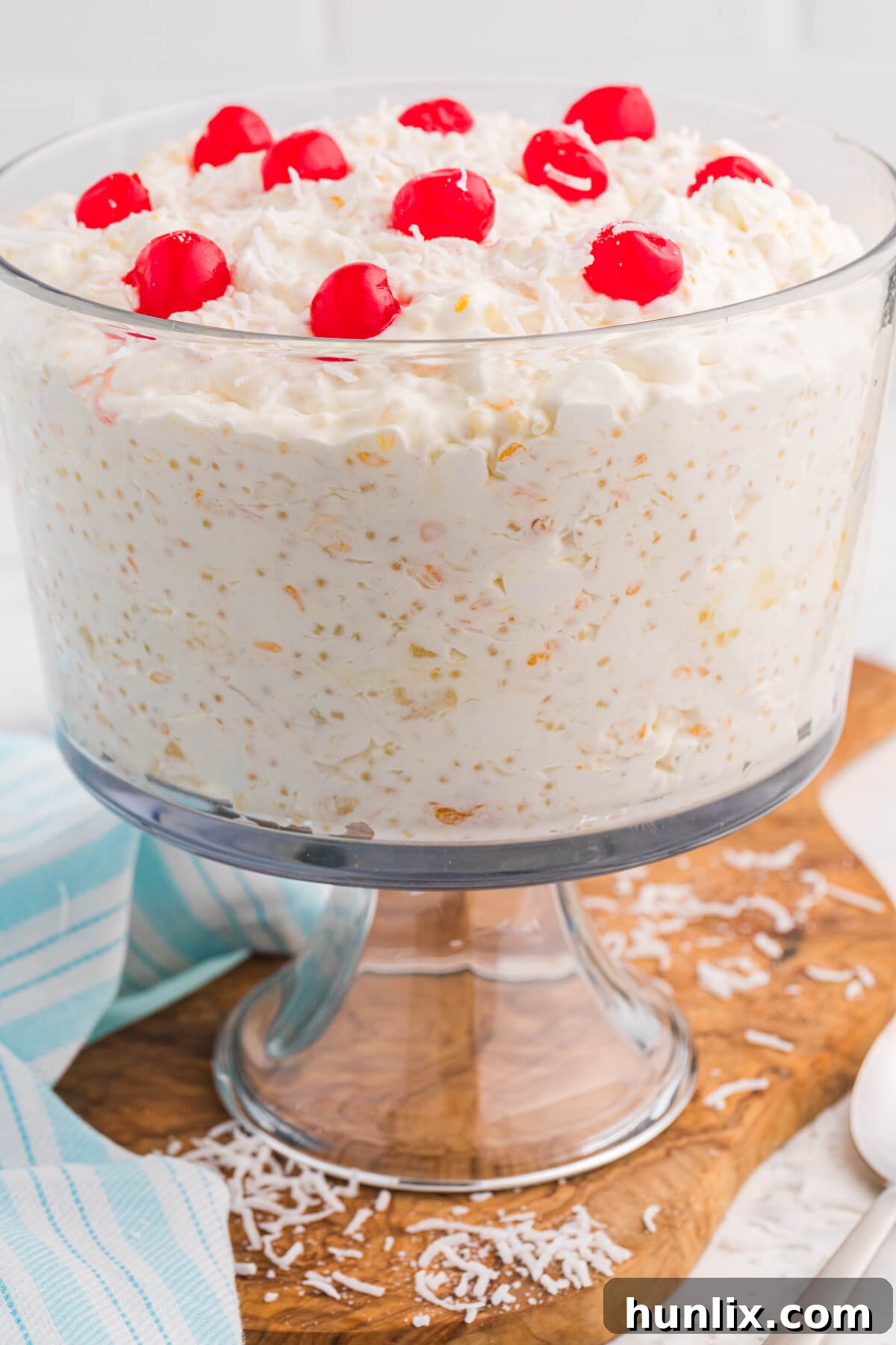 A beautifully presented Frog Eye Salad in a clear glass trifle bowl, adorned with a swirl of whipped cream, a sprinkle of shredded coconut, and bright red maraschino cherries, all placed on a rustic wooden board.