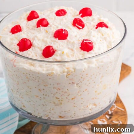 Creamy dessert salad in a clear glass bowl topped with whipped topping, shredded coconut flakes, and bright red maraschino cherries on a wooden serving board.