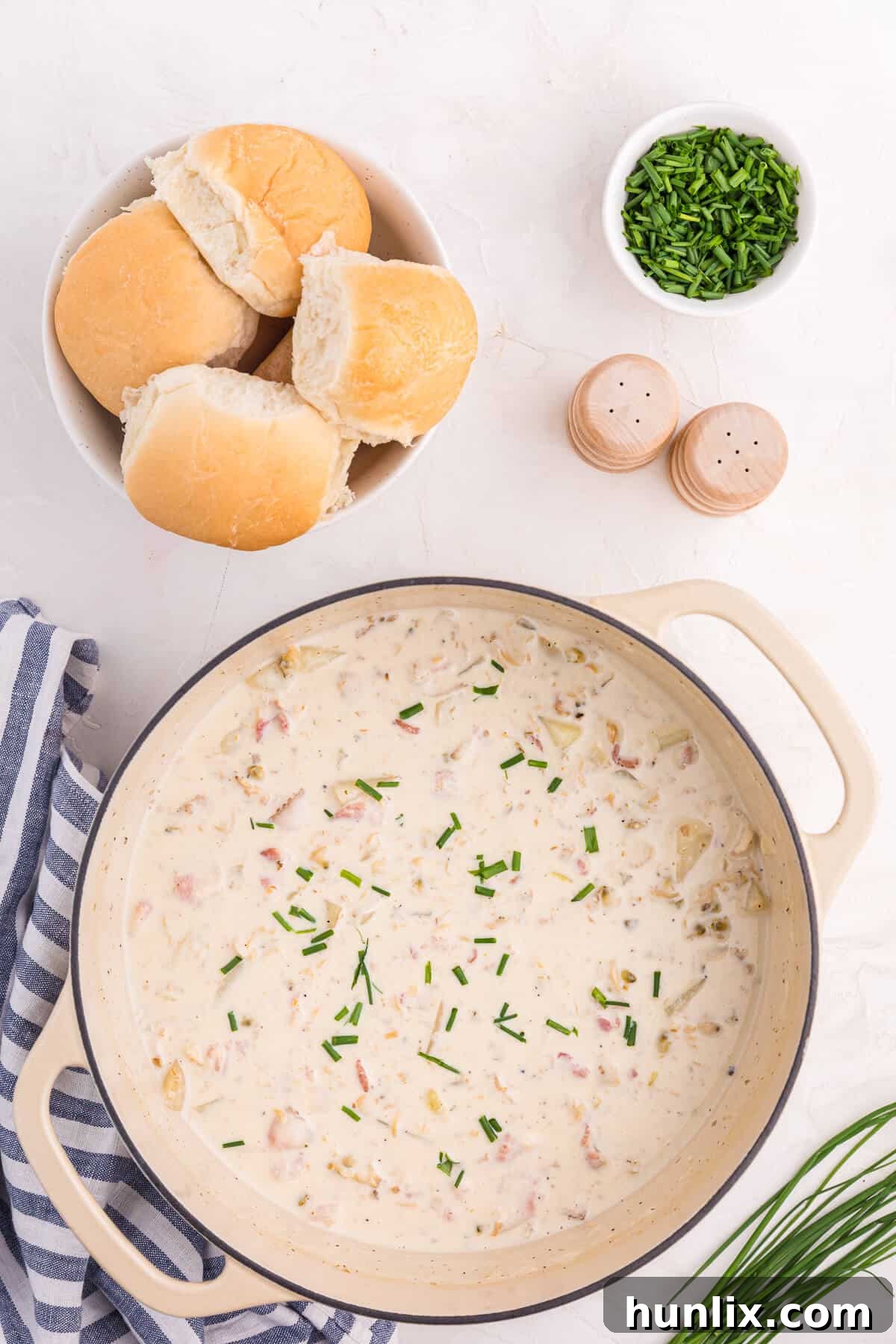 The Quintessential New England Clam Chowder 6 A large Dutch oven filled with New England Clam Chowder, served with dinner rolls and garnished with fresh chives, set on a rustic wooden surface.