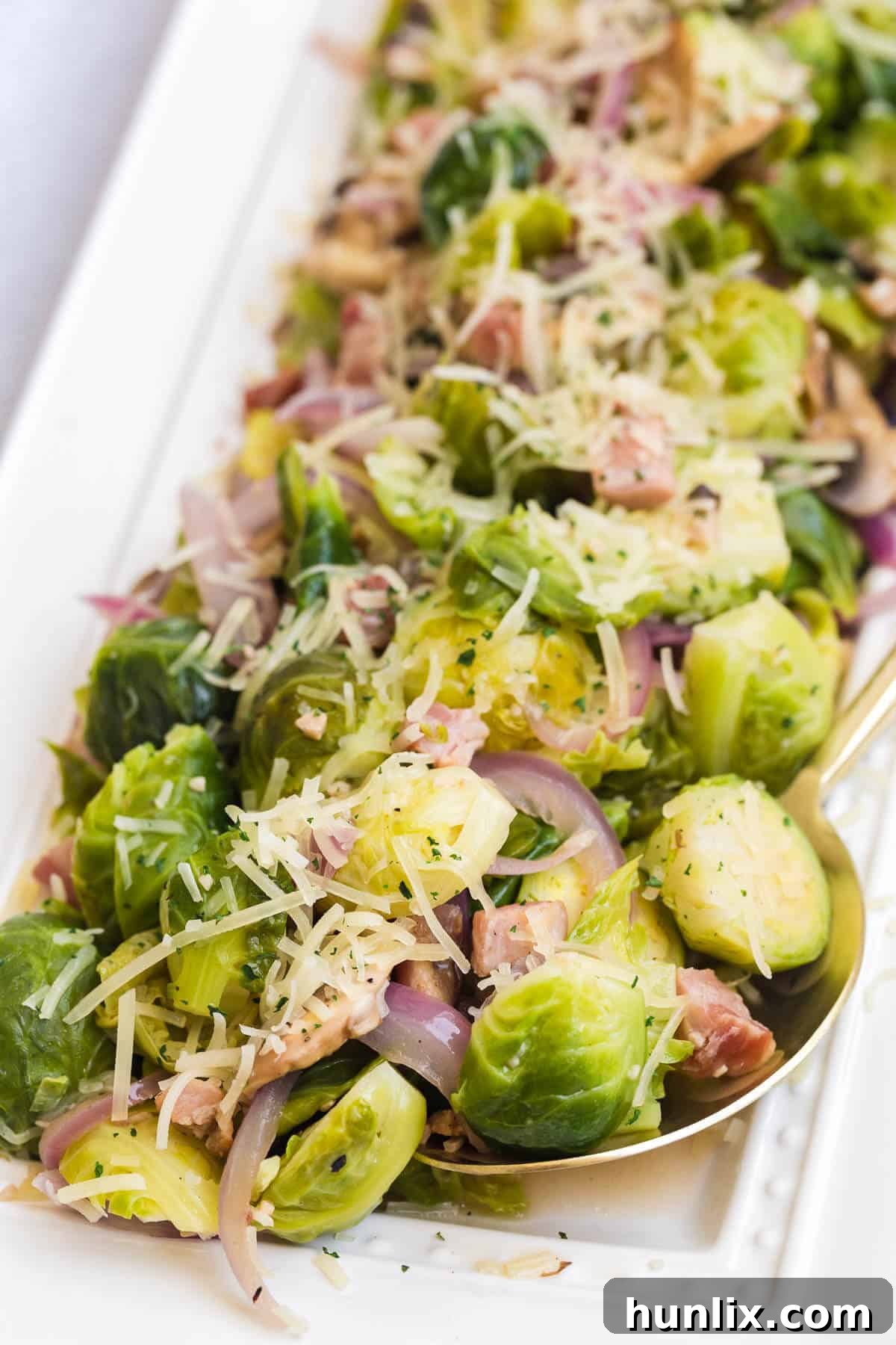 Close-up of a serving spoon scooping perfectly cooked Brussels sprouts with lemon, crispy pancetta, and sautéed mushrooms from a large white platter.