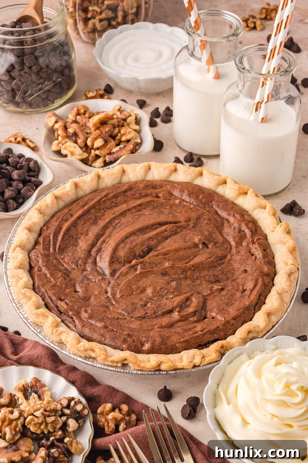 A beautifully baked Brownie Pie, still in its pie dish, surrounded by various ingredients used in its creation like chocolate chips, walnuts, and butter, on a wooden surface.