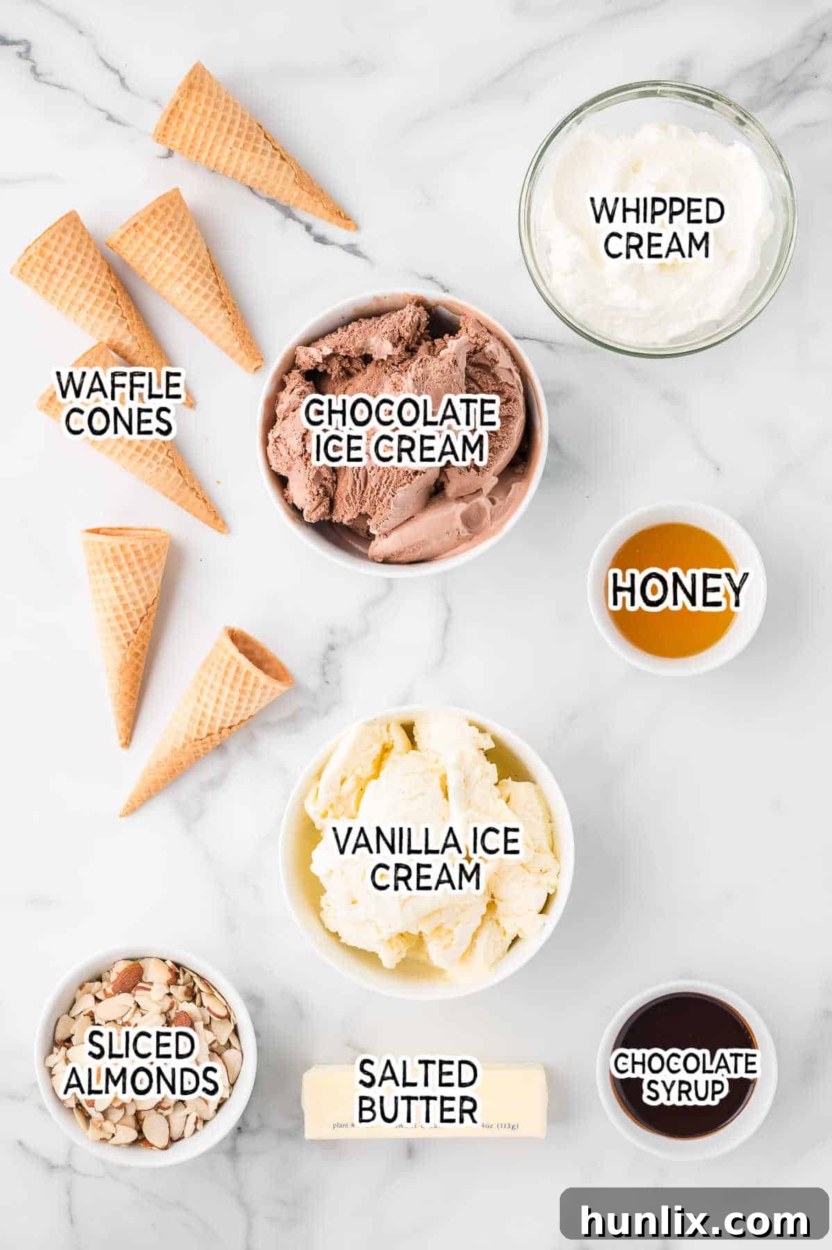 A collection of fresh ingredients for making ice cream pie, including honey, butter, waffle cones, sliced almonds, vanilla and chocolate ice cream, and chocolate syrup, all neatly arranged on a white marble counter.