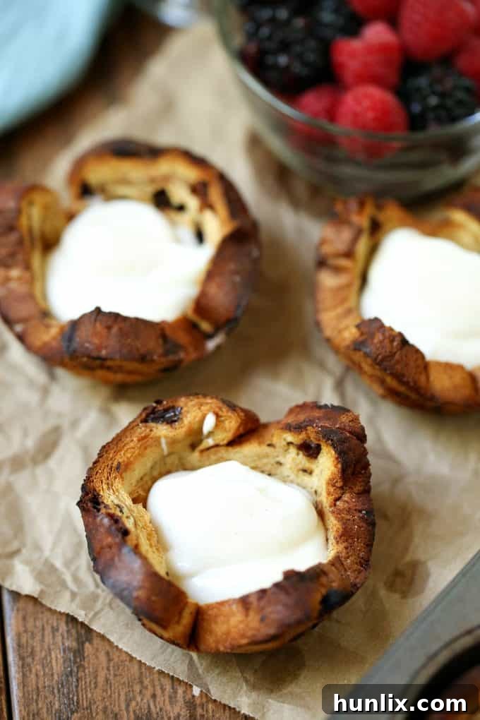 Two baked cinnamon raisin bread cups, one filled with Greek yogurt and mixed berries, and the other awaiting toppings.