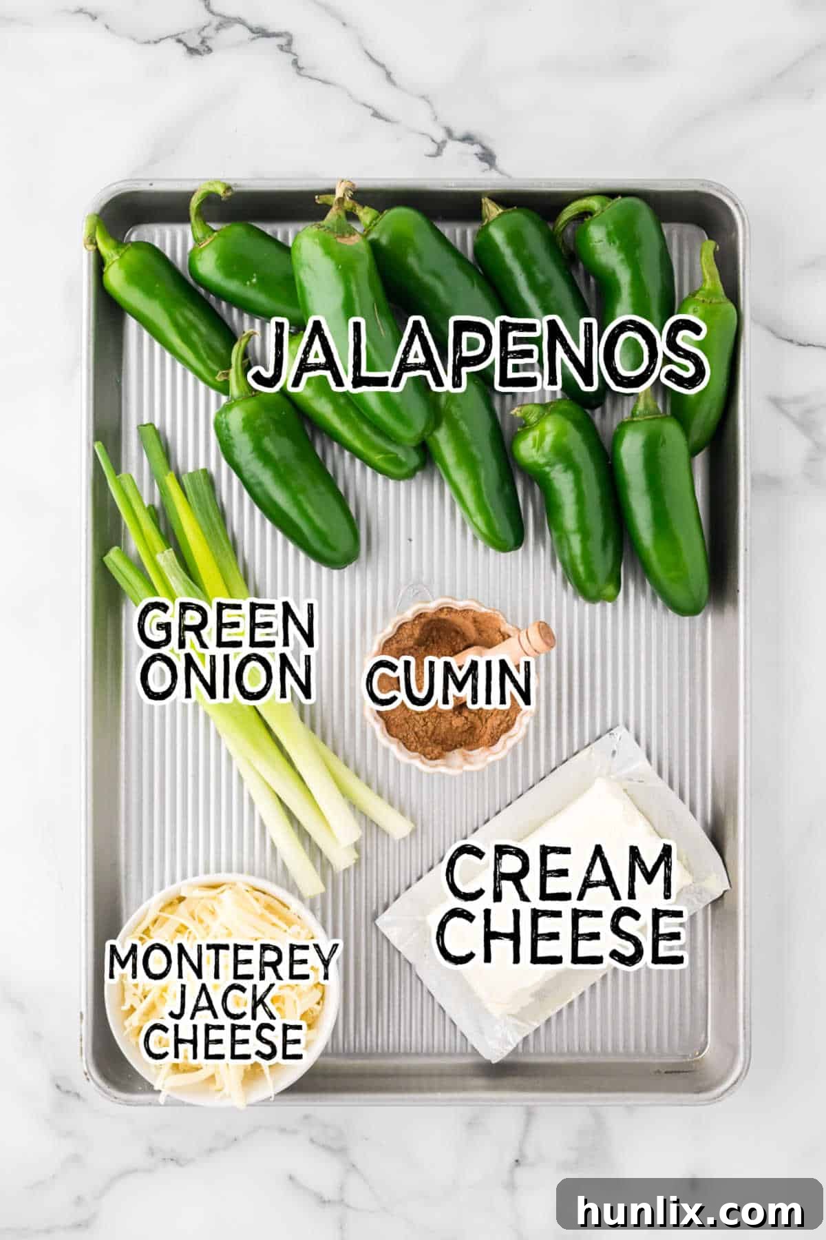 Various fresh ingredients laid out on a cutting board, including whole jalapenos, a block of cream cheese, a bowl of shredded cheese, cumin, and chopped green onions.