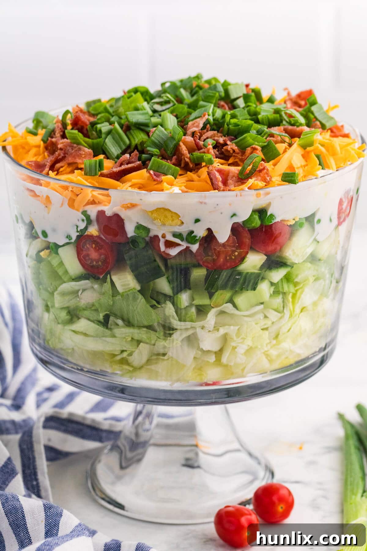 Seven layer salad served in a pedestal glass bowl with visible vegetable layers, creamy dressing, cheddar cheese, bacon, and green onion topping.
