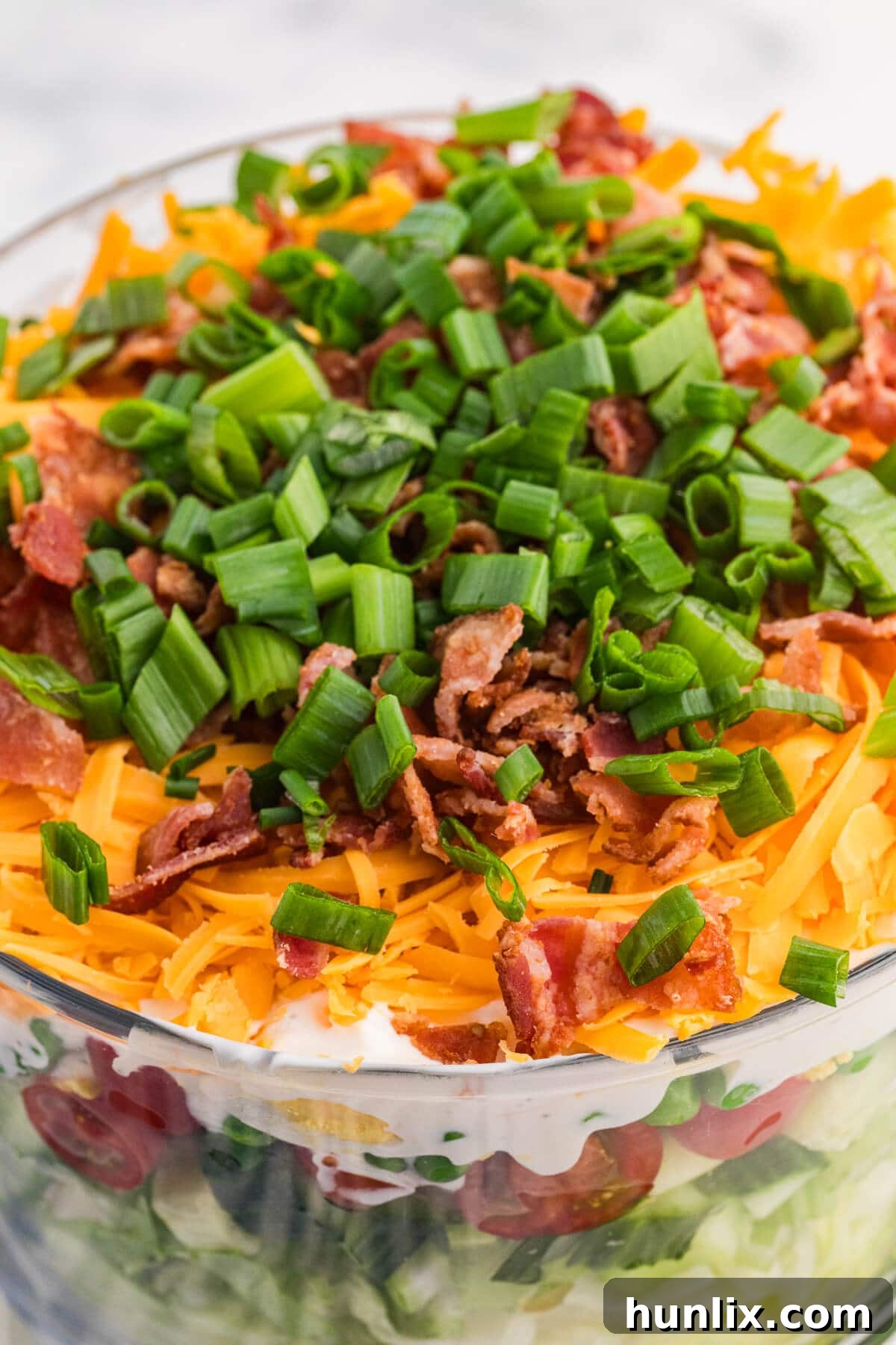 Close-up of seven layer salad topped with shredded cheddar cheese, crispy bacon, and fresh green onions in a glass serving bowl.